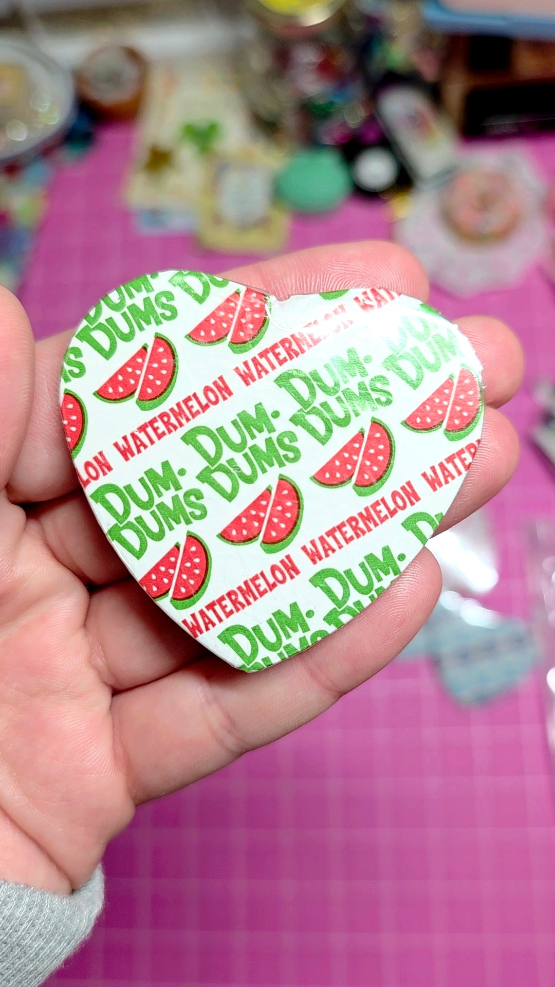 Heart-shaped Dum-Dum's watermelon water candy wrapper held in a hand with a pink background.