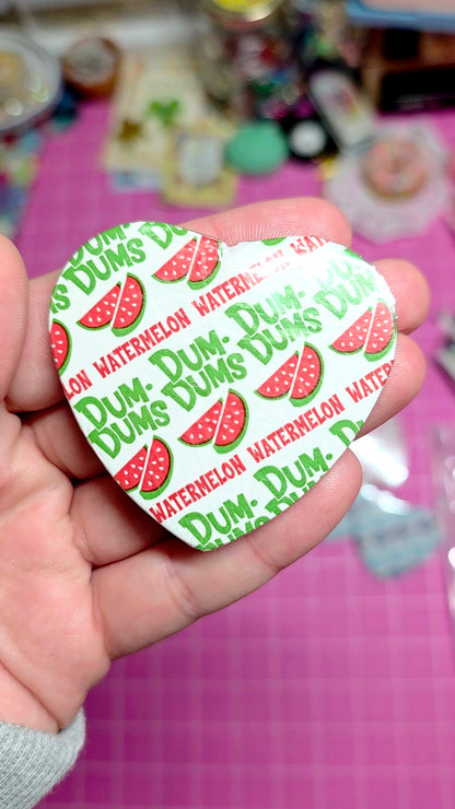 Heart-shaped Dum-Dum's watermelon water candy wrapper held in a hand with a pink background.