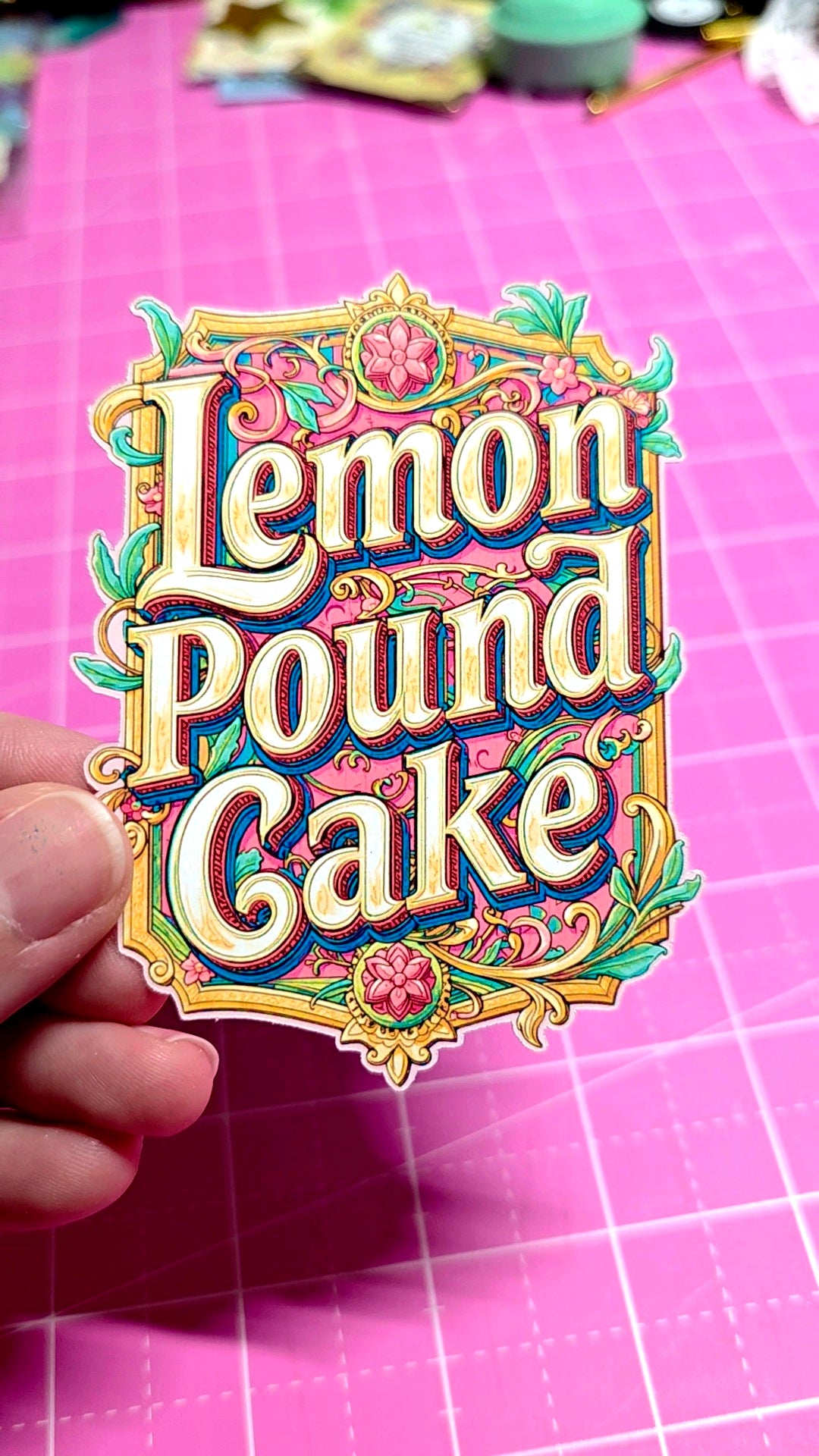 Decorative sticker with 'Lemon Pound Cake' text held over a pink cutting mat.