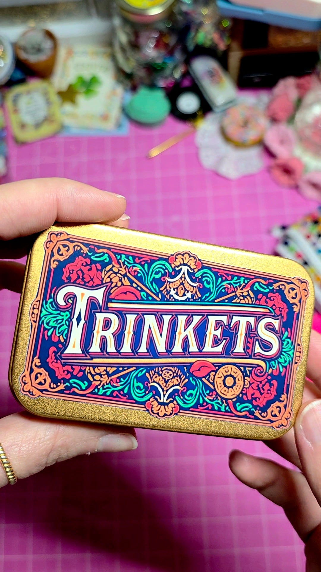 Decorative tin labeled 'Trinkets' held by a hand on a pink surface with various items in the background.