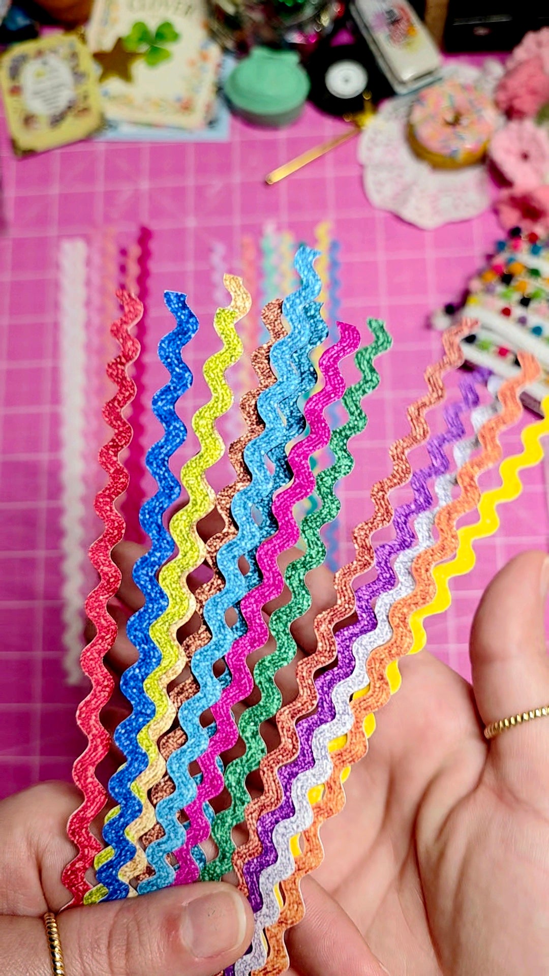Colorful zigzag-patterned paper strips held by a hand on a pink surface with decorative items.