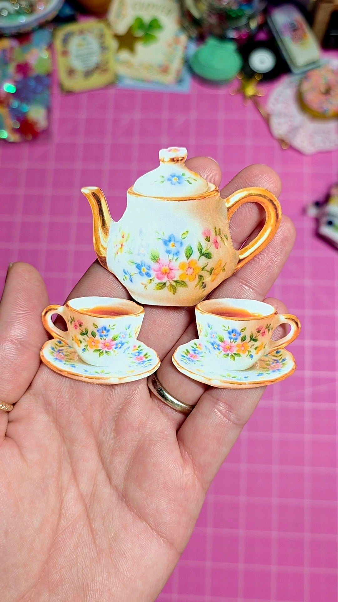 Miniature teapot and cups with floral design held in a hand against a pink background