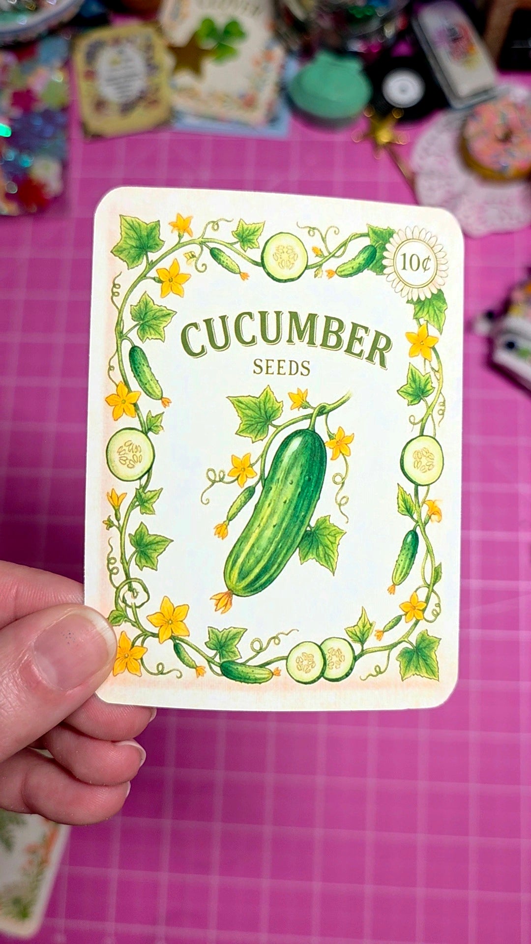 Card with cucumber seeds packaging held by a hand on a pink grid background
