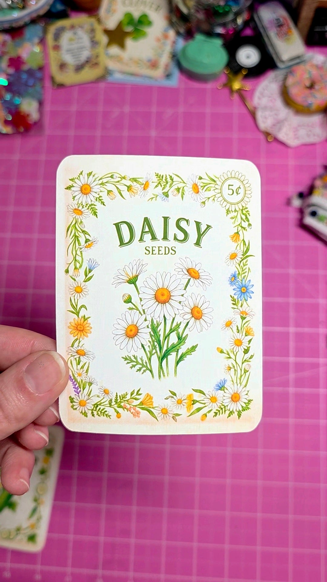 Card with 'Daisy Seeds' design held over a pink grid background