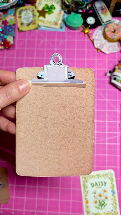 Brown clipboard held by a hand on a pink cutting mat with various items in the background.