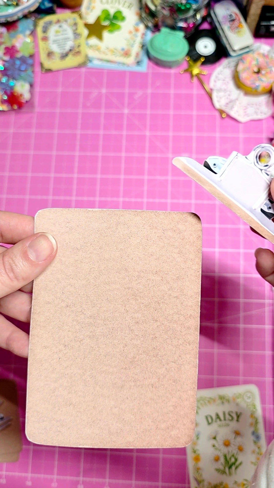 Hand holding a beige card against a pink grid background with decorative items.