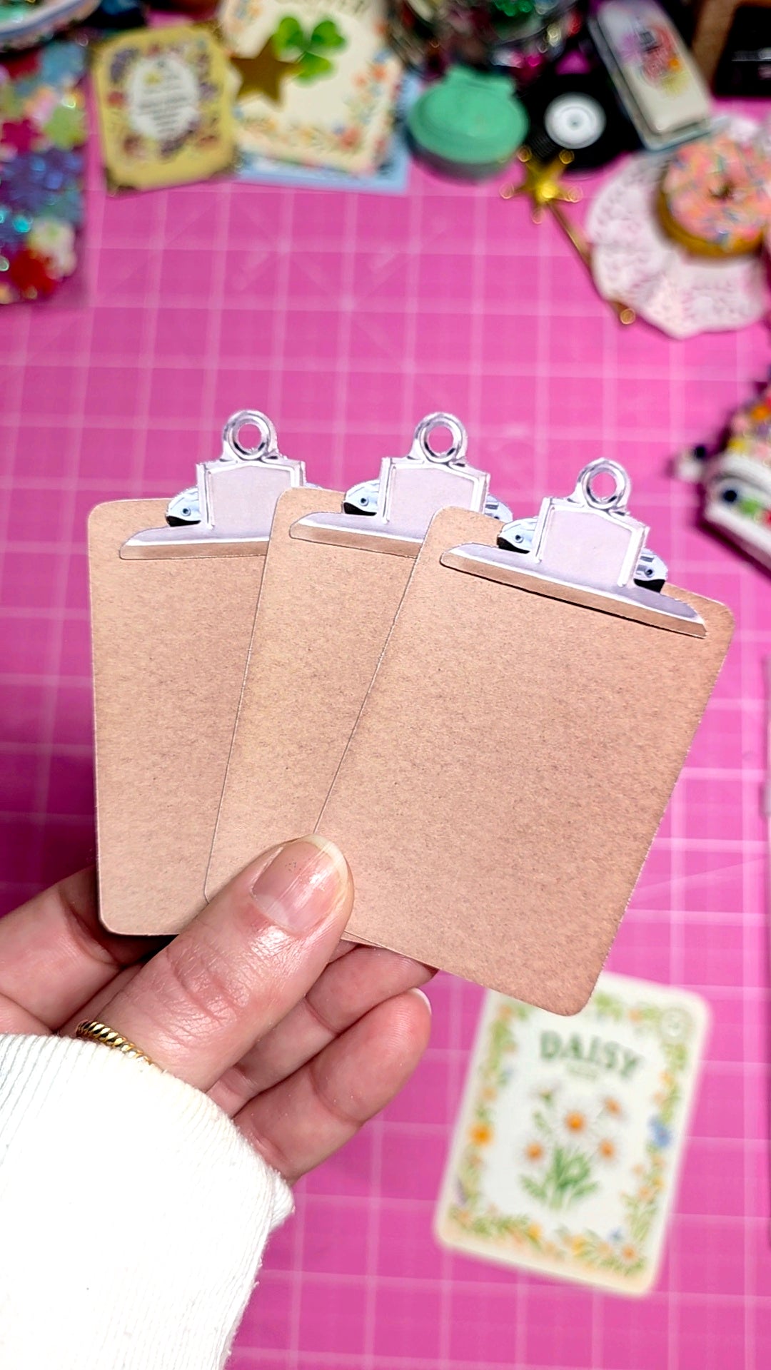 Three small brown clipboards held by a hand on a pink surface with colorful items in the background.