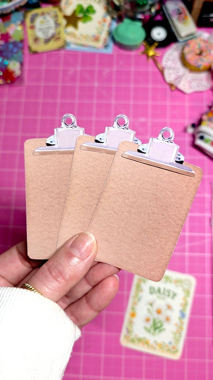 Three small brown clipboards held by a hand on a pink surface with colorful items in the background.