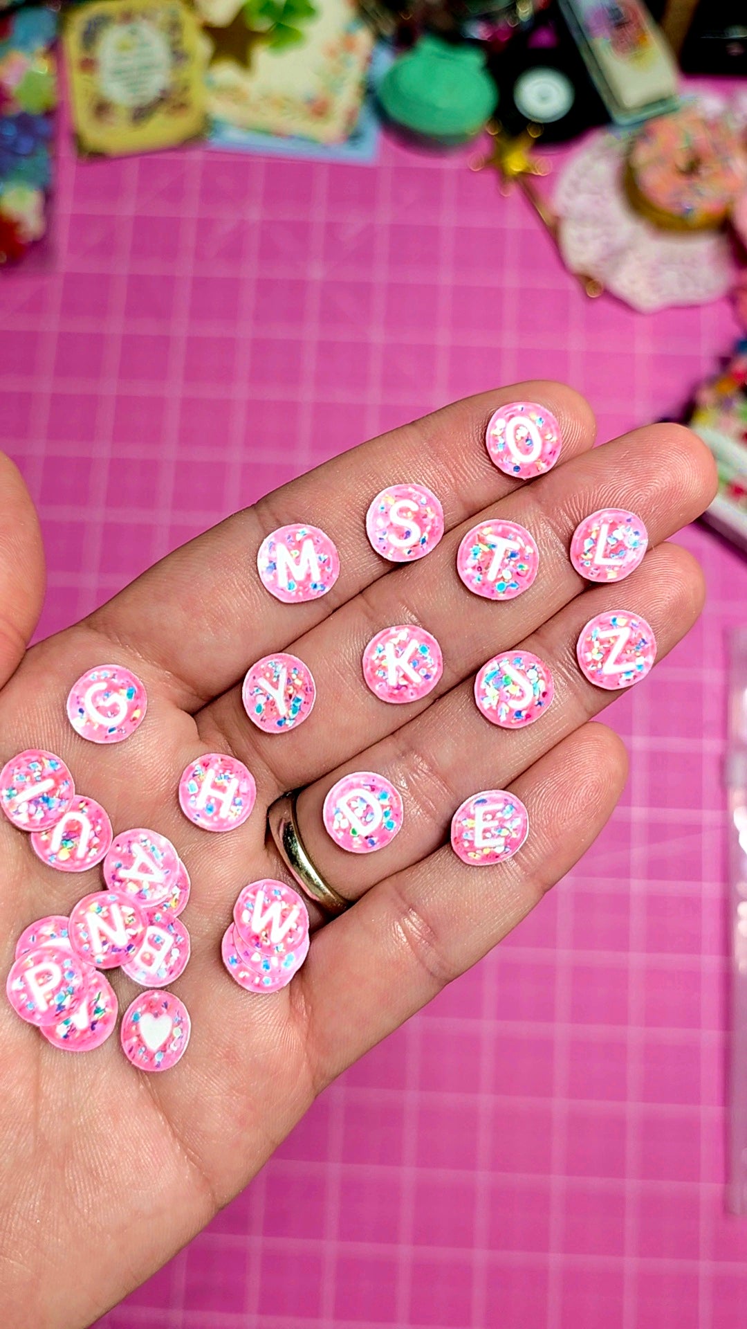 Hand holding pink decorative buttons with letters and designs on a pink checkered background