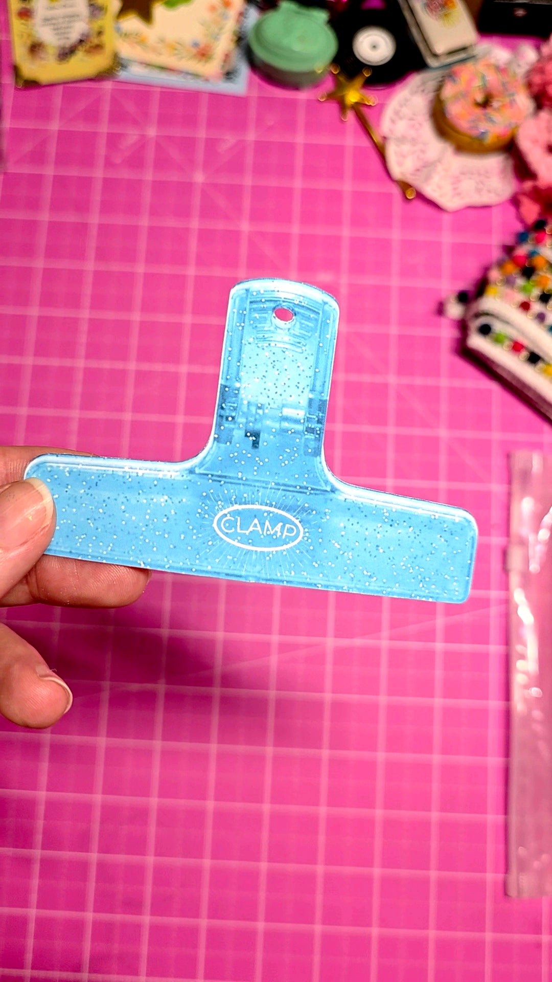 Blue paper clip held over a pink cutting mat with various items in the background