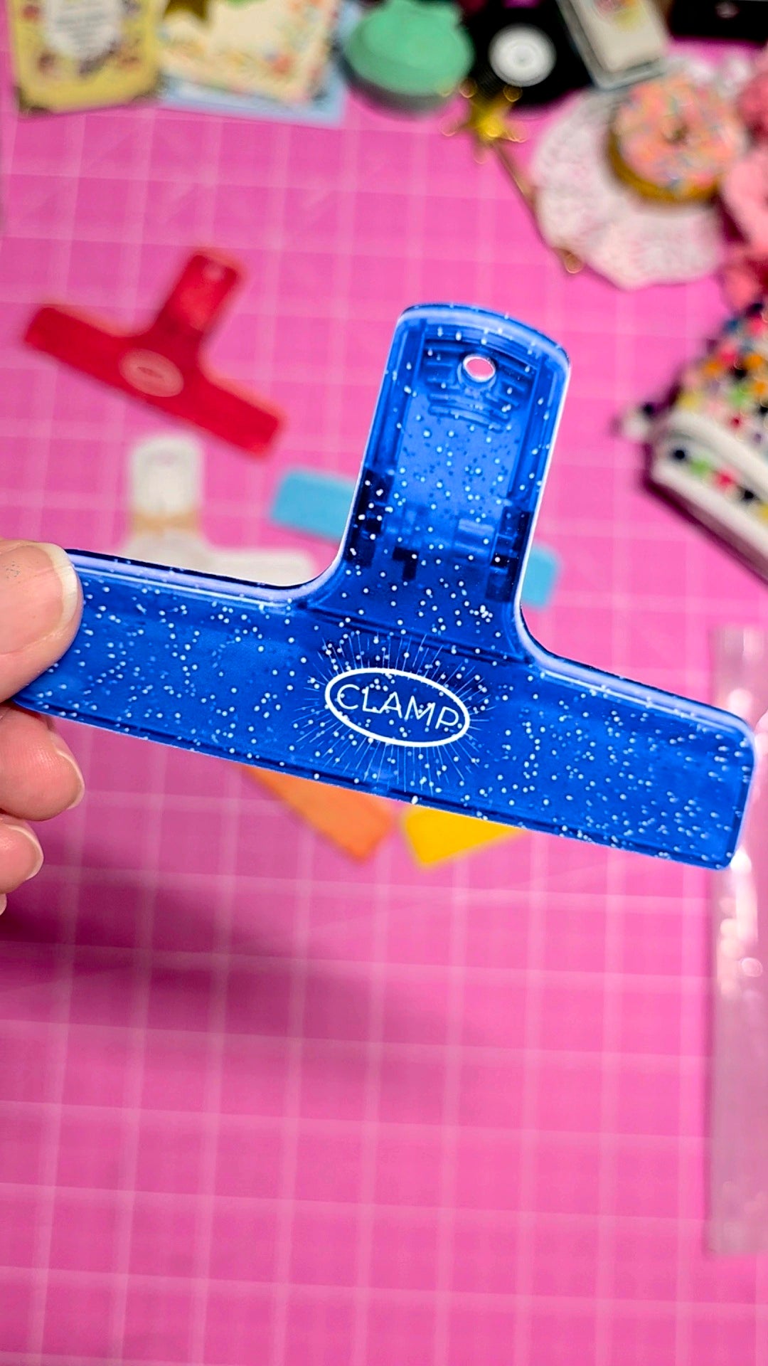 Blue paper clip with 'CLAMA' branding held against a pink background with colorful objects.