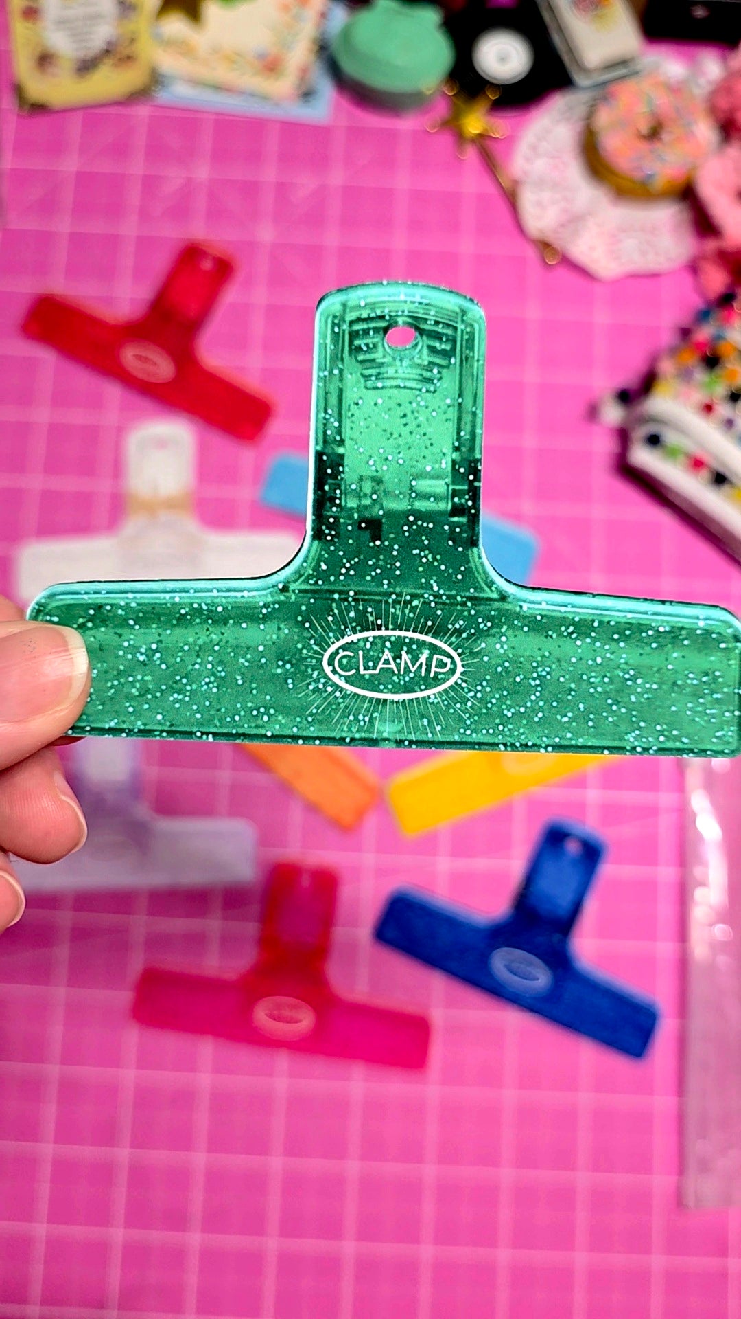 Green paper clip with 'CLAMA' branding held over a pink cutting mat with colorful paper clips.