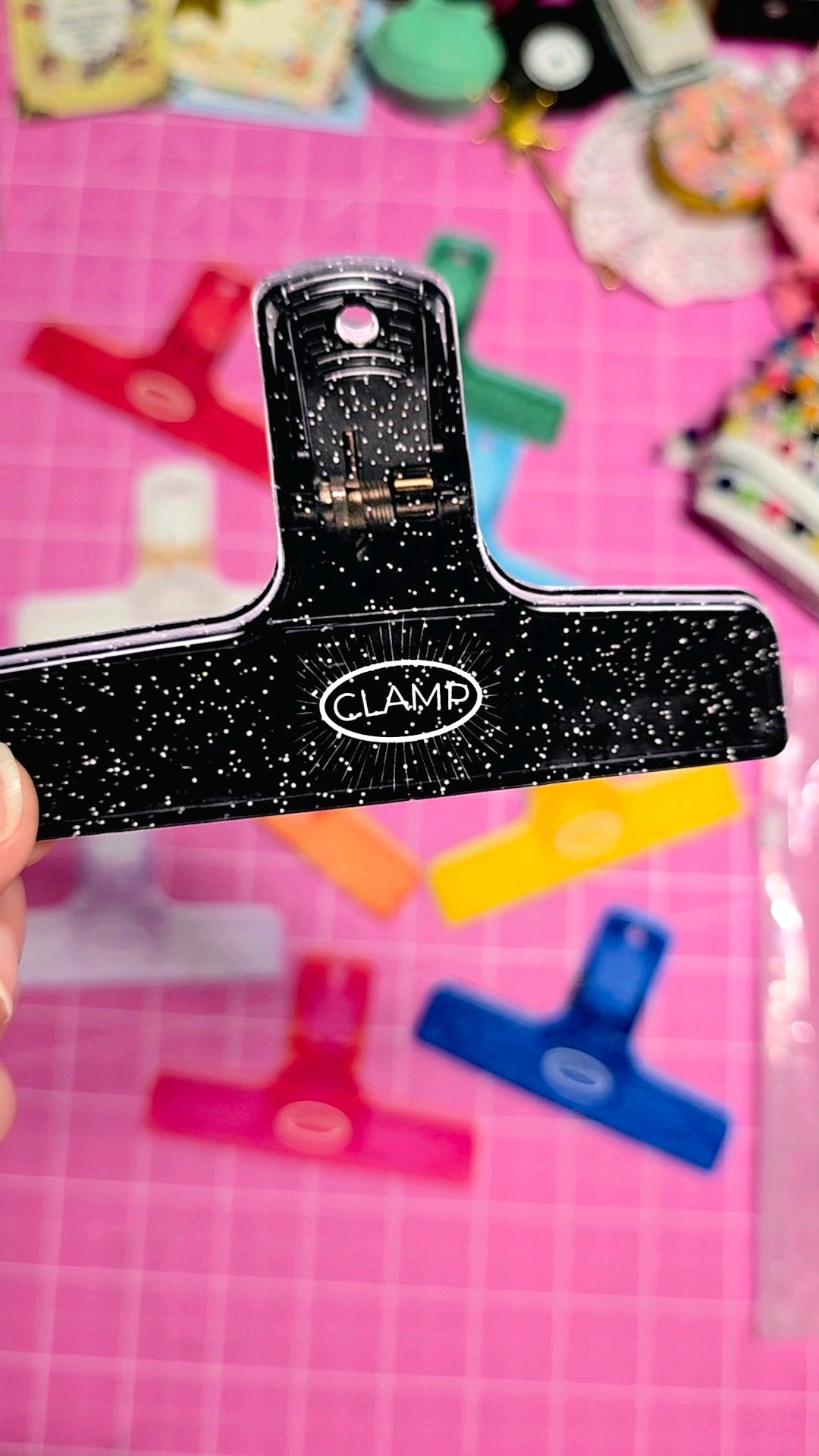 Black paper clip with 'CLAMP' branding on a pink background with colorful objects.