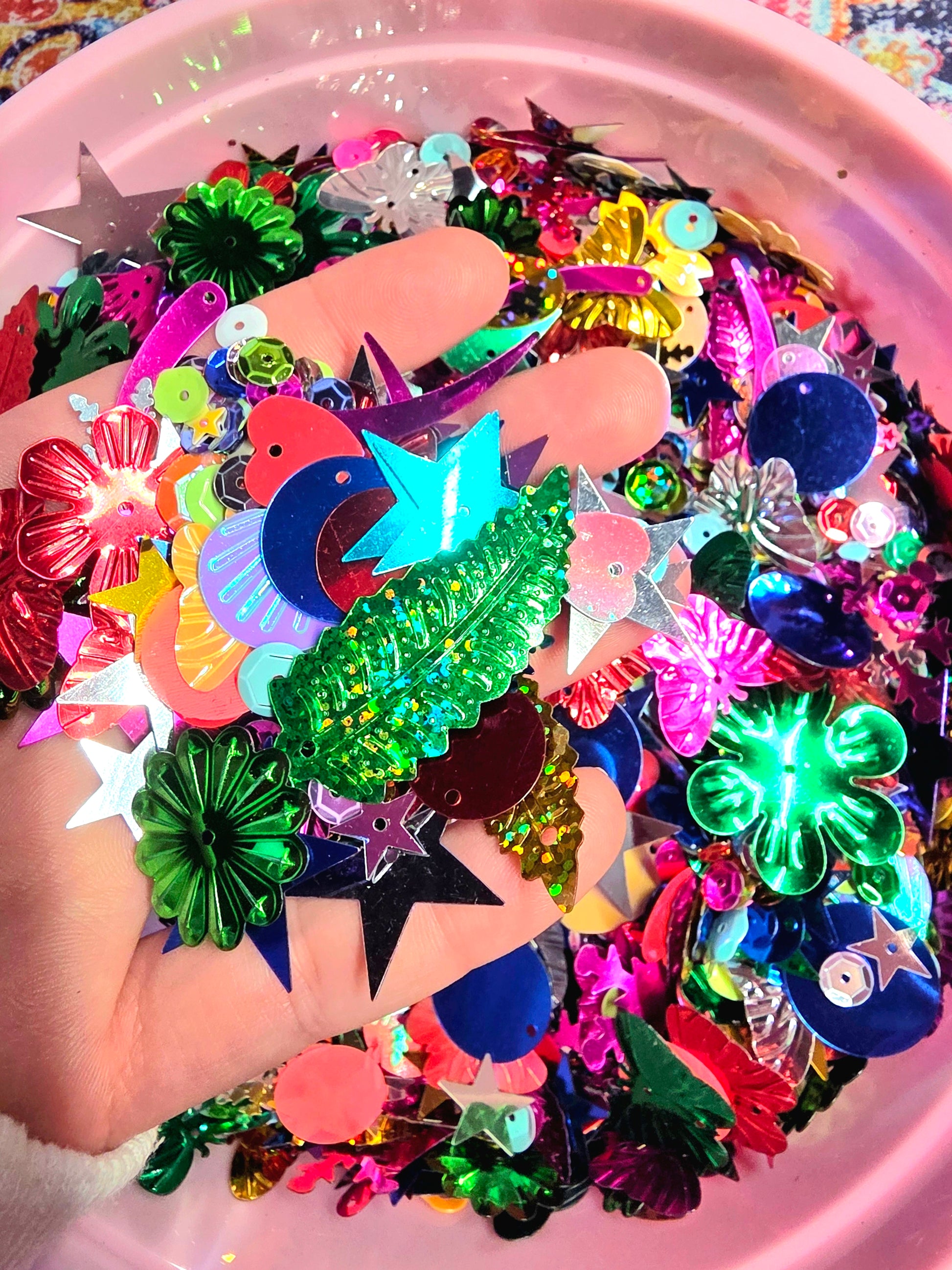 Colorful plastic embellishments held by a hand with a pink container filled with more embellishments in the background.