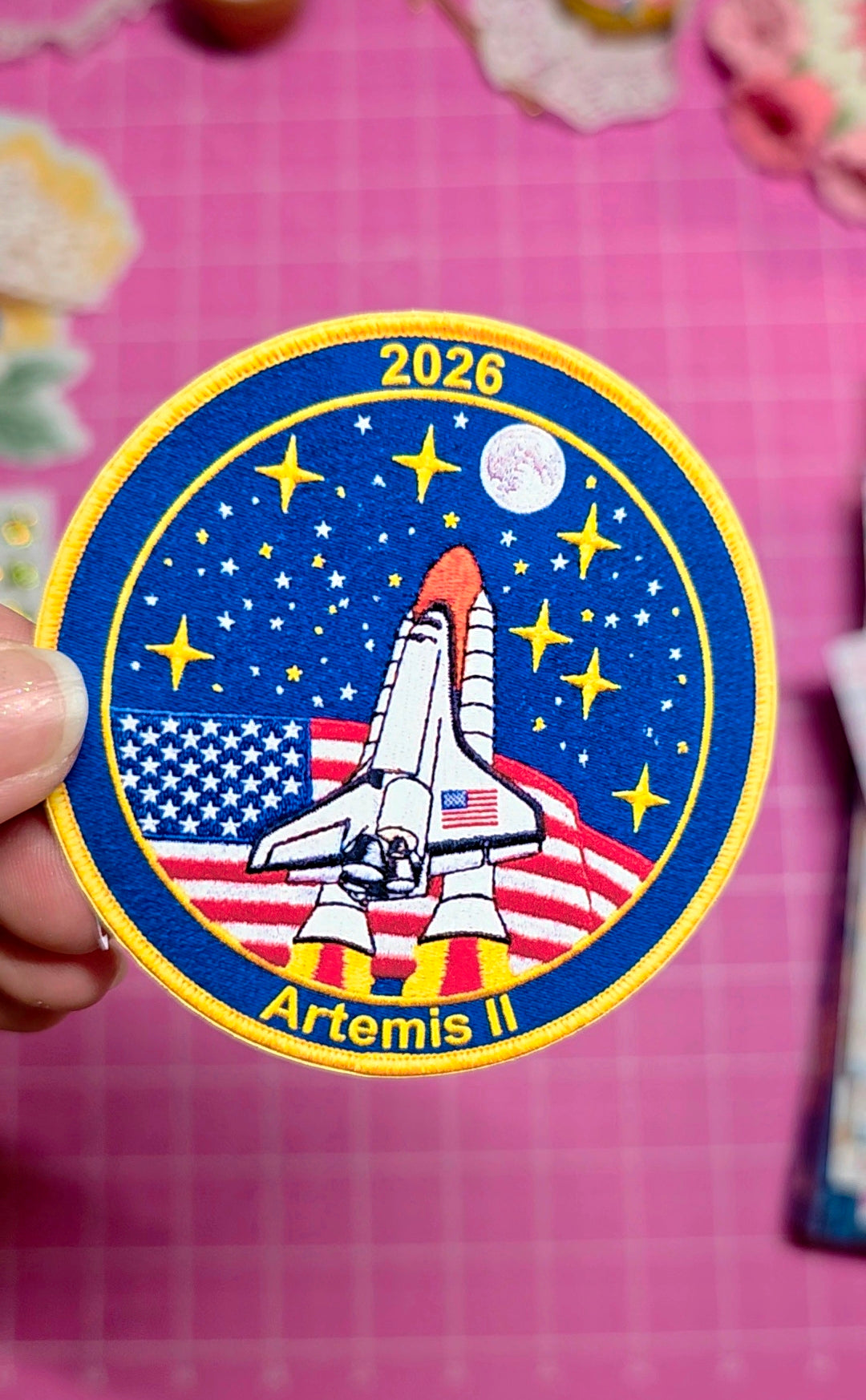 Space-themed patch with space shuttle, stars, and American flag on a pink background