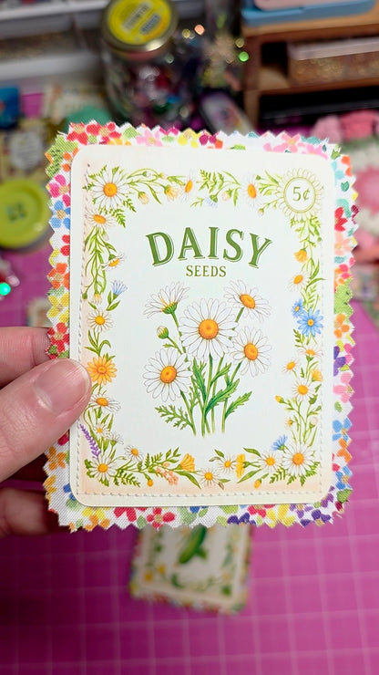 Card with 'Daisy Seeds' design held by a hand on a pink surface.