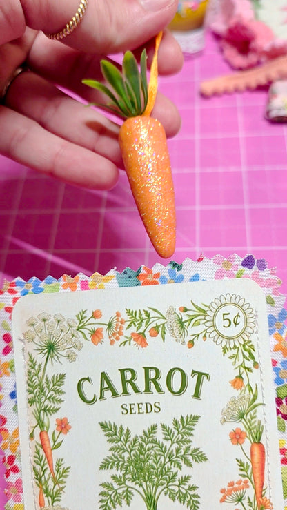 Hand holding a decorative carrot next to a seed packet labeled 'Carrot Seeds' on a pink checkered background.