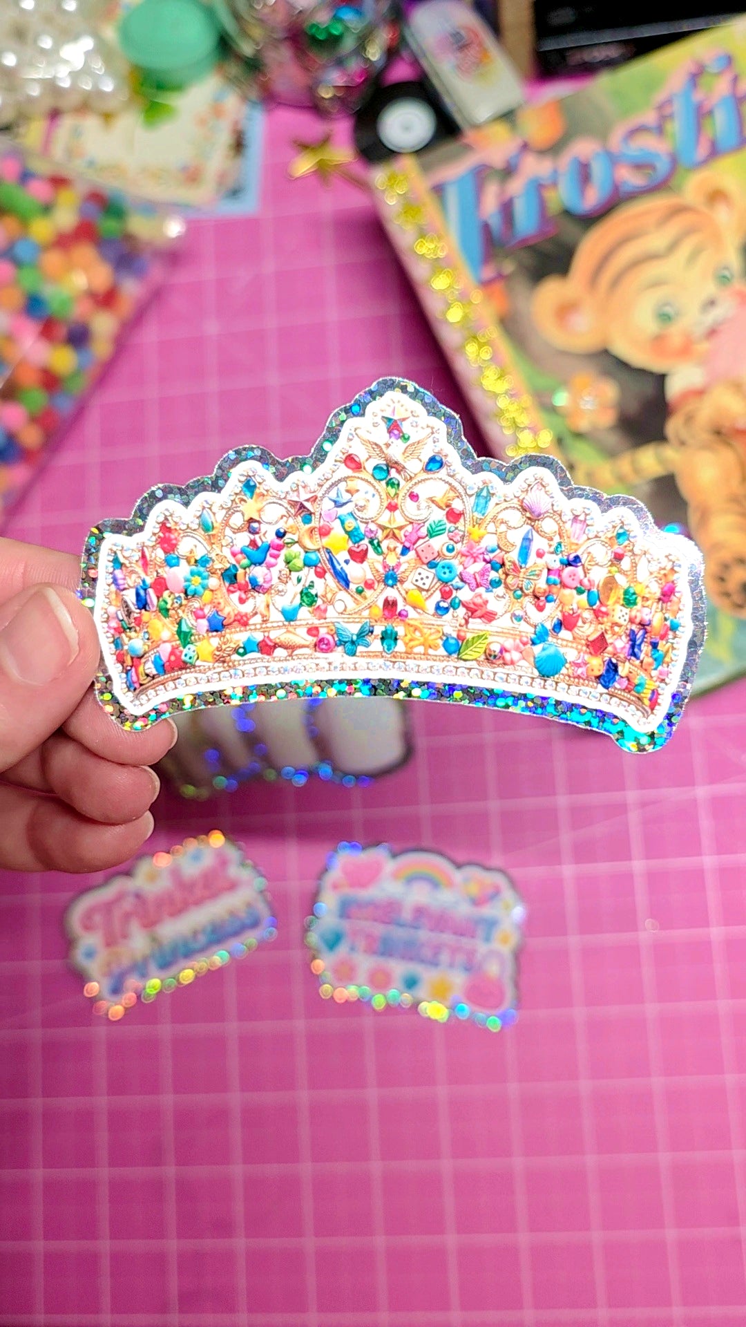Colorful crown-shaped sticker held over a pink surface with scattered stickers.