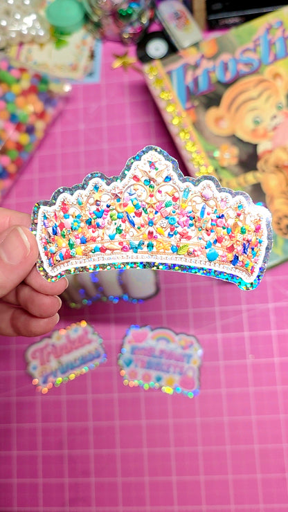 Colorful crown-shaped sticker held over a pink surface with scattered stickers.