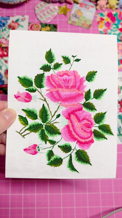 Embroidered fabric with pink roses on a white card held over a colorful fabric background.