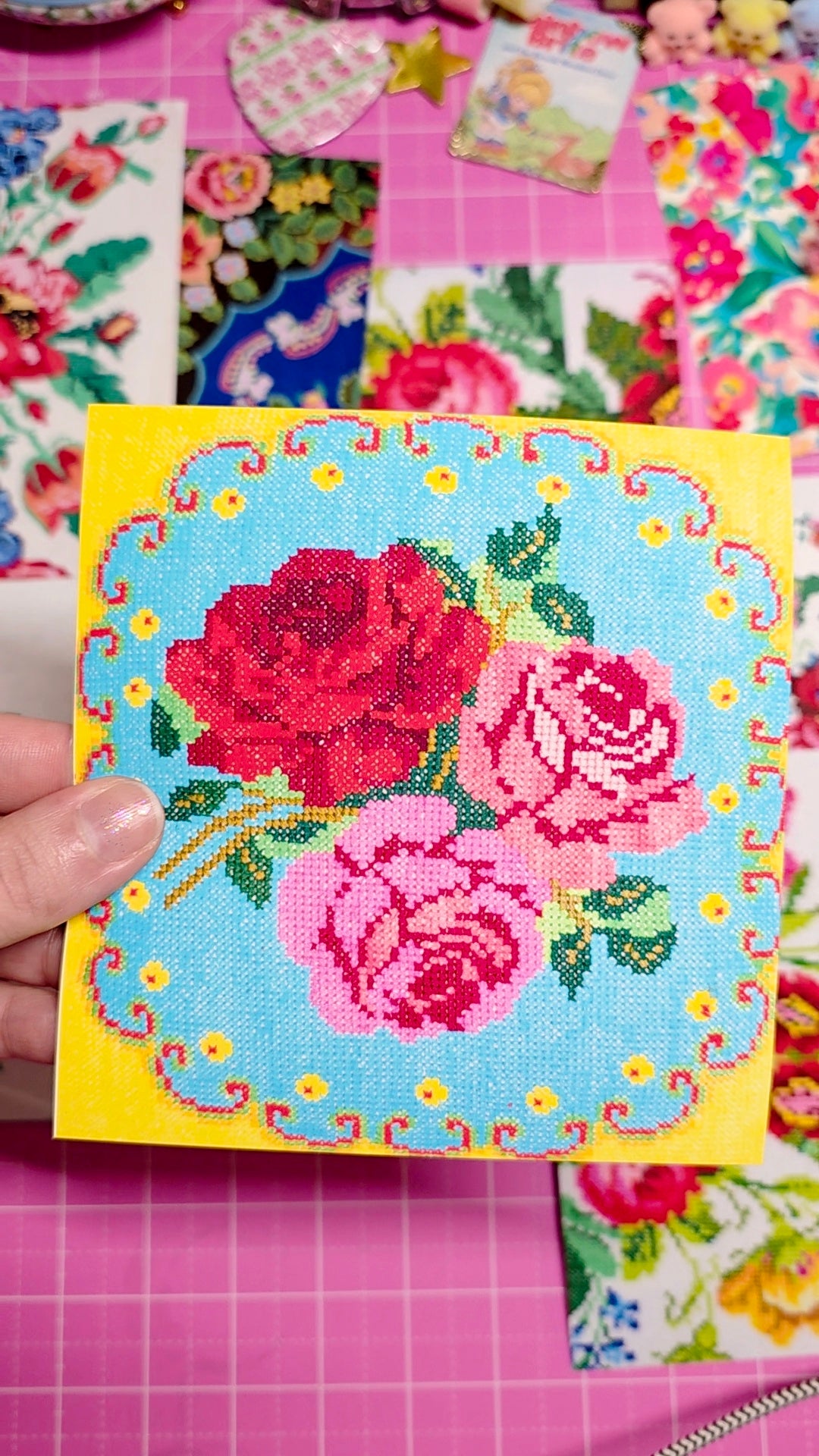 Cross-stitched rose design on a yellow card with a pink floral background