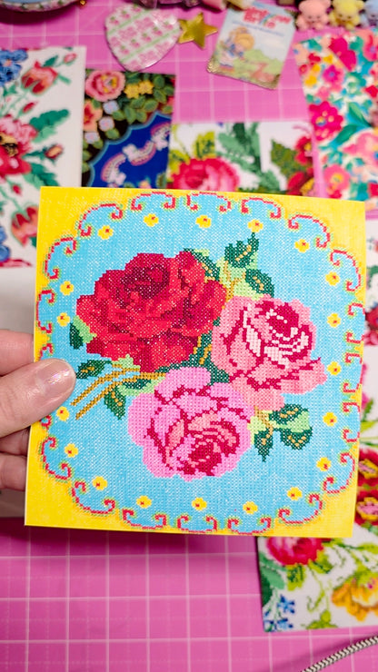 Cross-stitched rose design on a yellow card with a pink floral background
