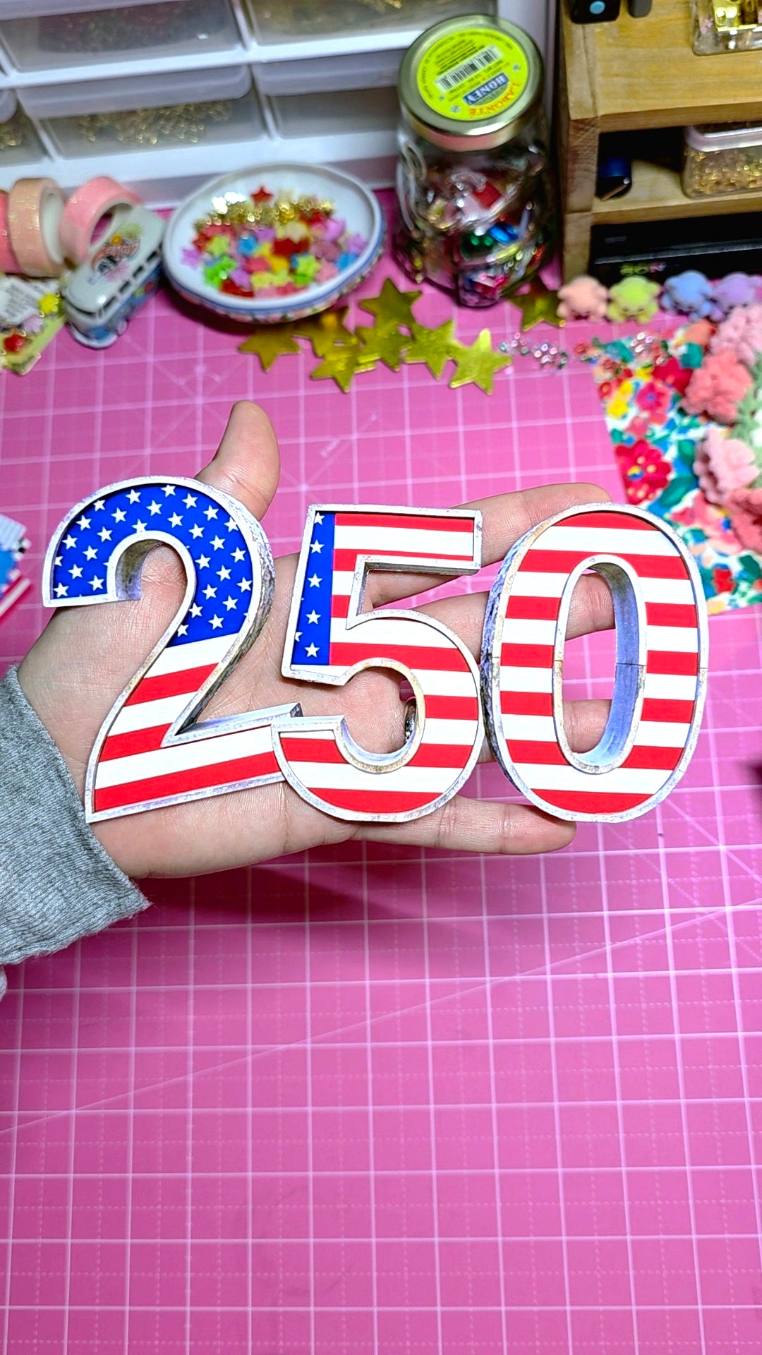 Hand holding decorative numbers with American flag design on a pink cutting mat.