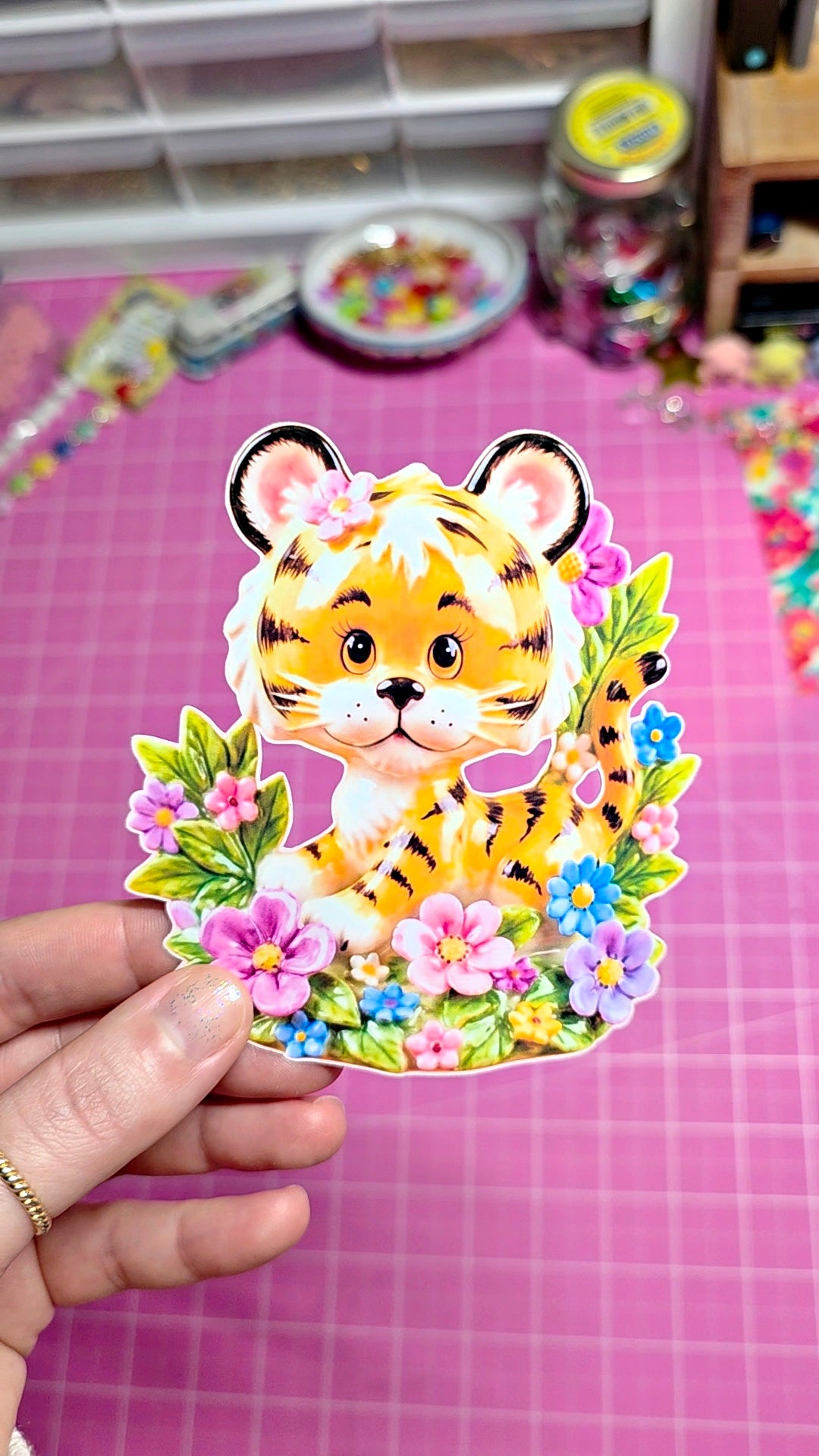 Sticker of a tiger surrounded by flowers held over a pink surface with crafting materials.