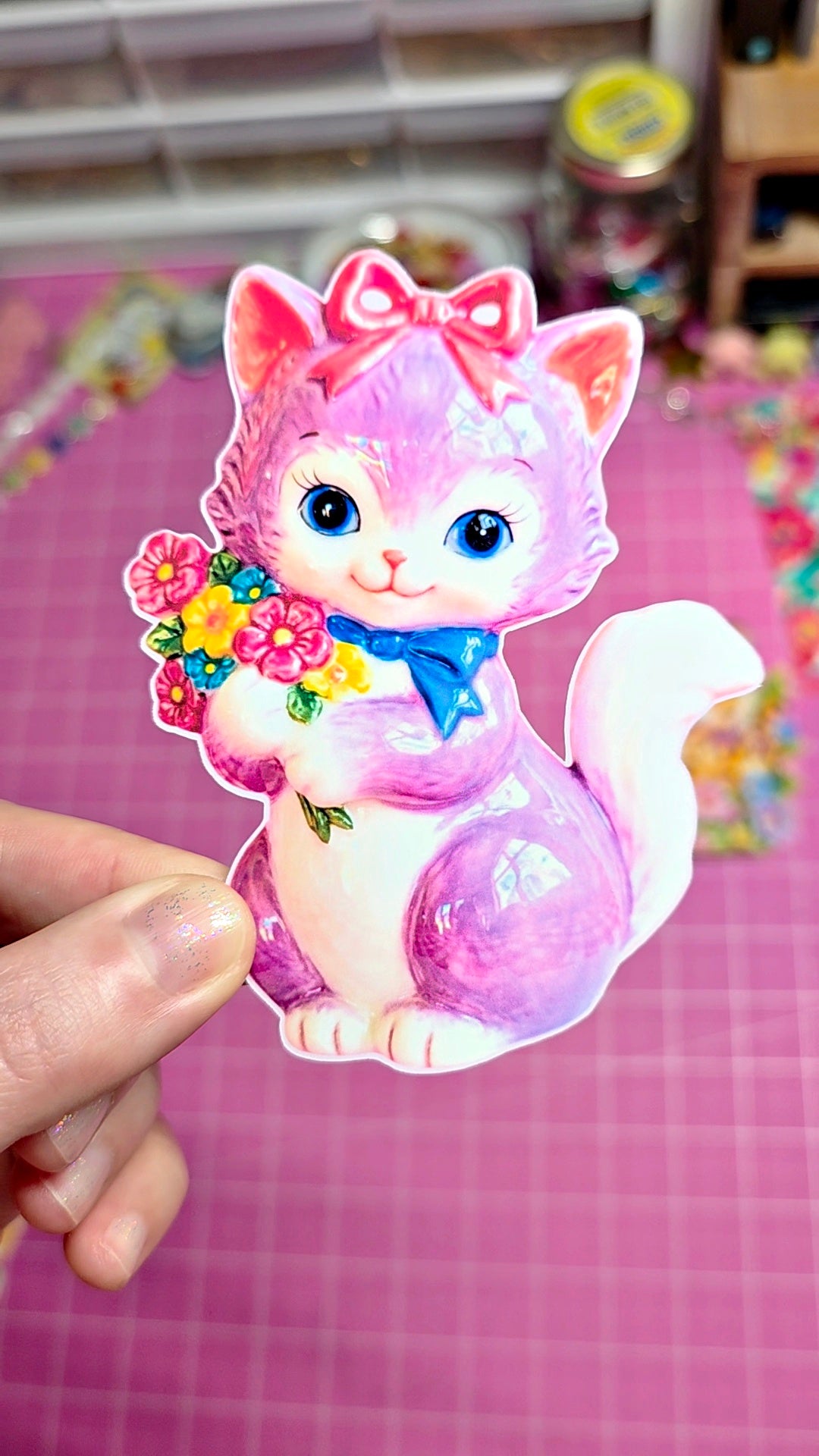 Colorful cartoon cat sticker held by a hand on a pink checkered background