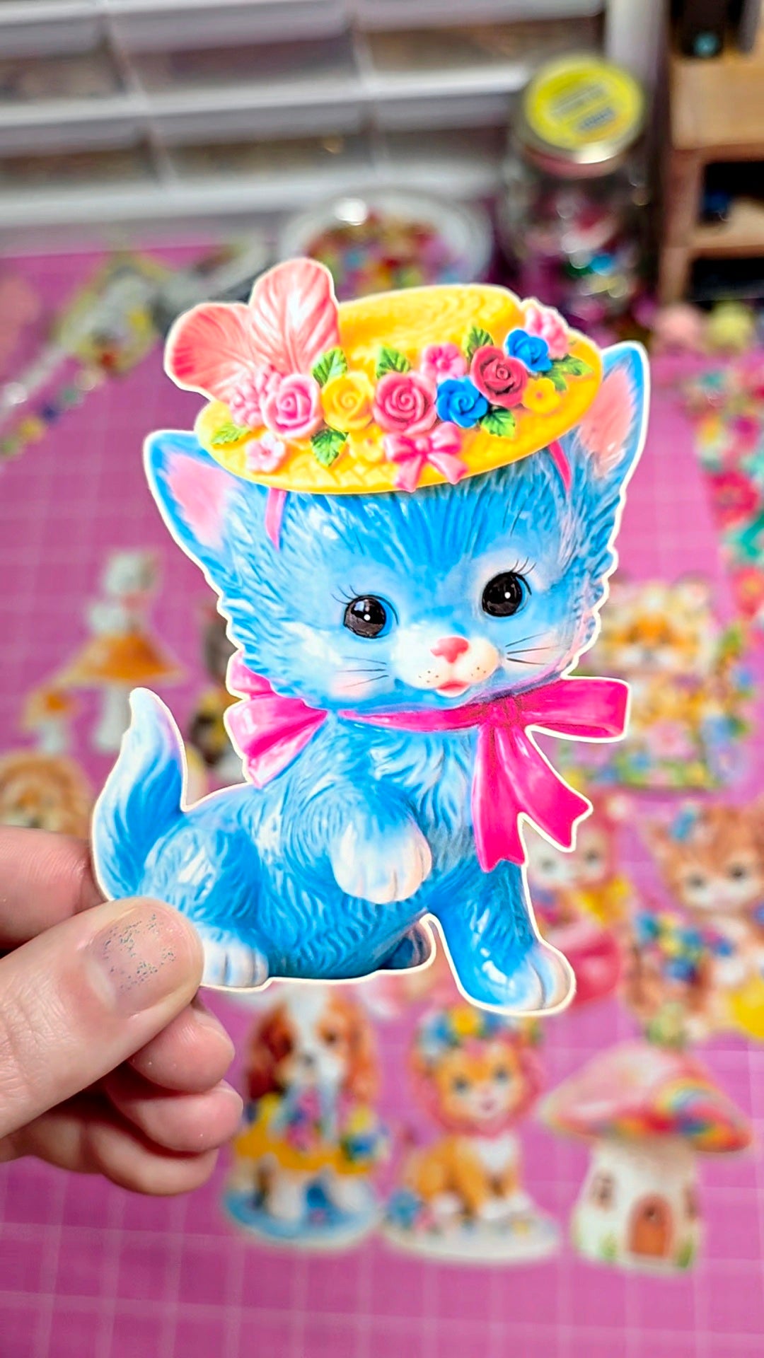 Blue cat figurine with a colorful hat held by a hand on a pink background with more cat figurines.