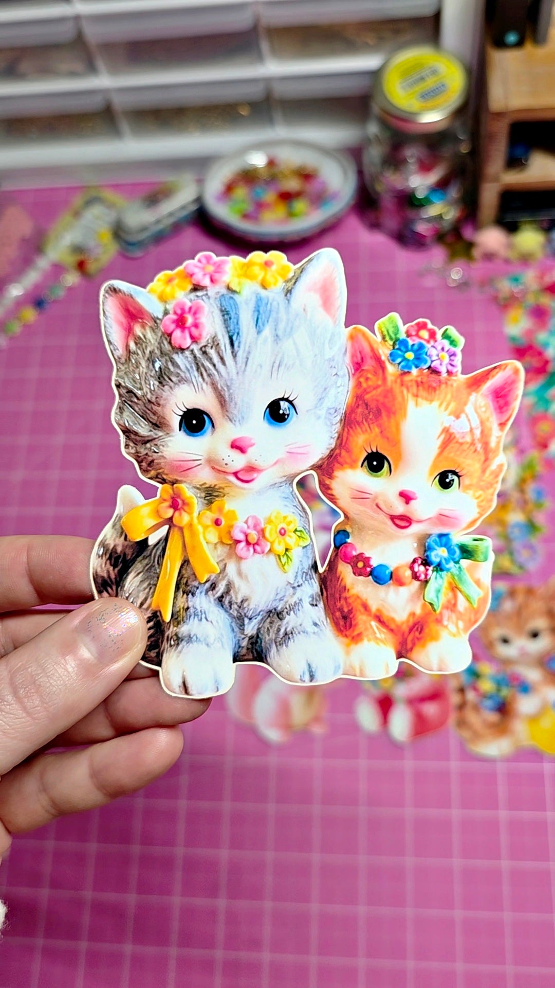 Two colorful cat stickers held by a hand on a pink grid background.