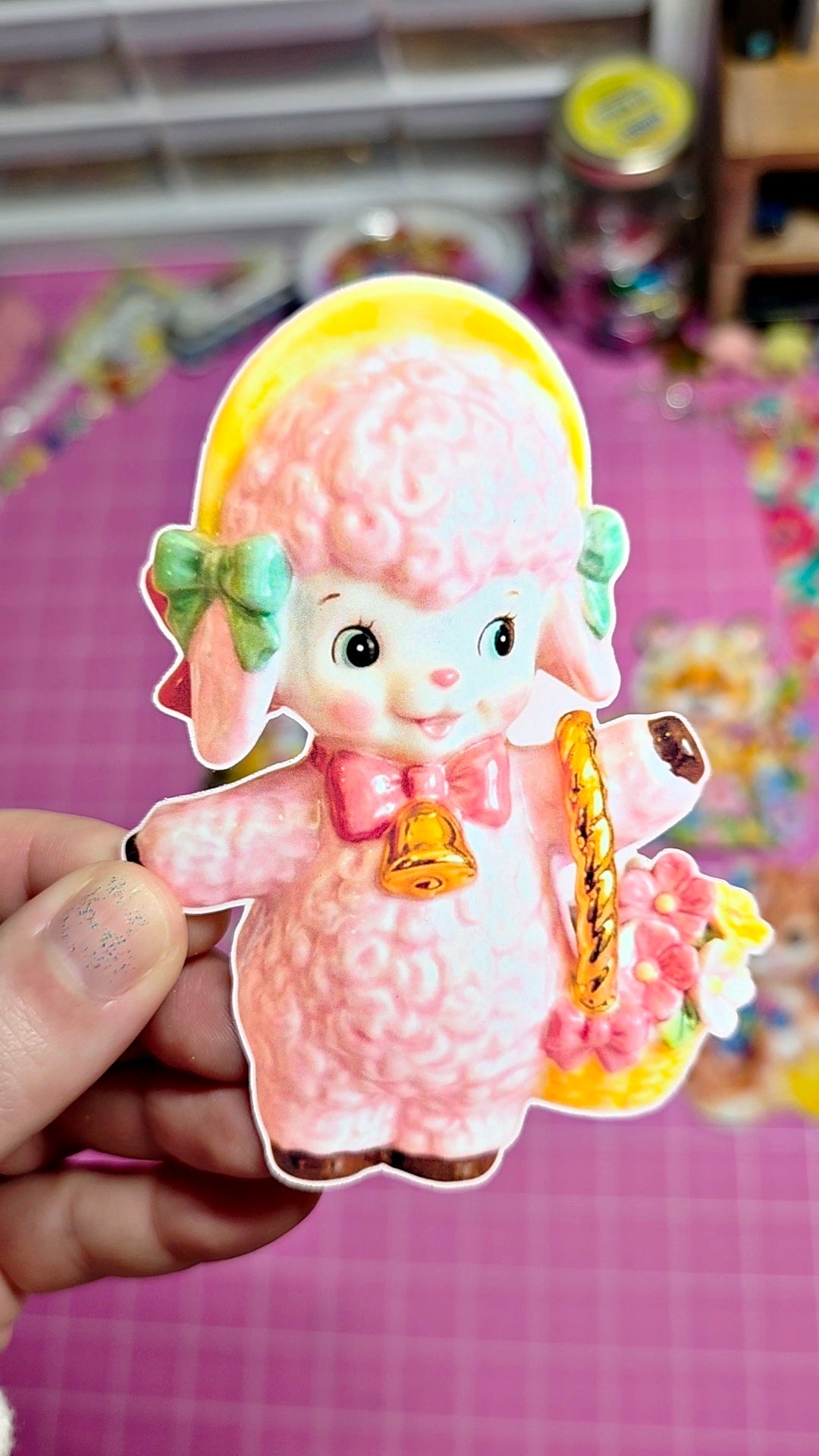 Cartoon lamb character with a yellow bow and pink dress held by a hand on a colorful background.