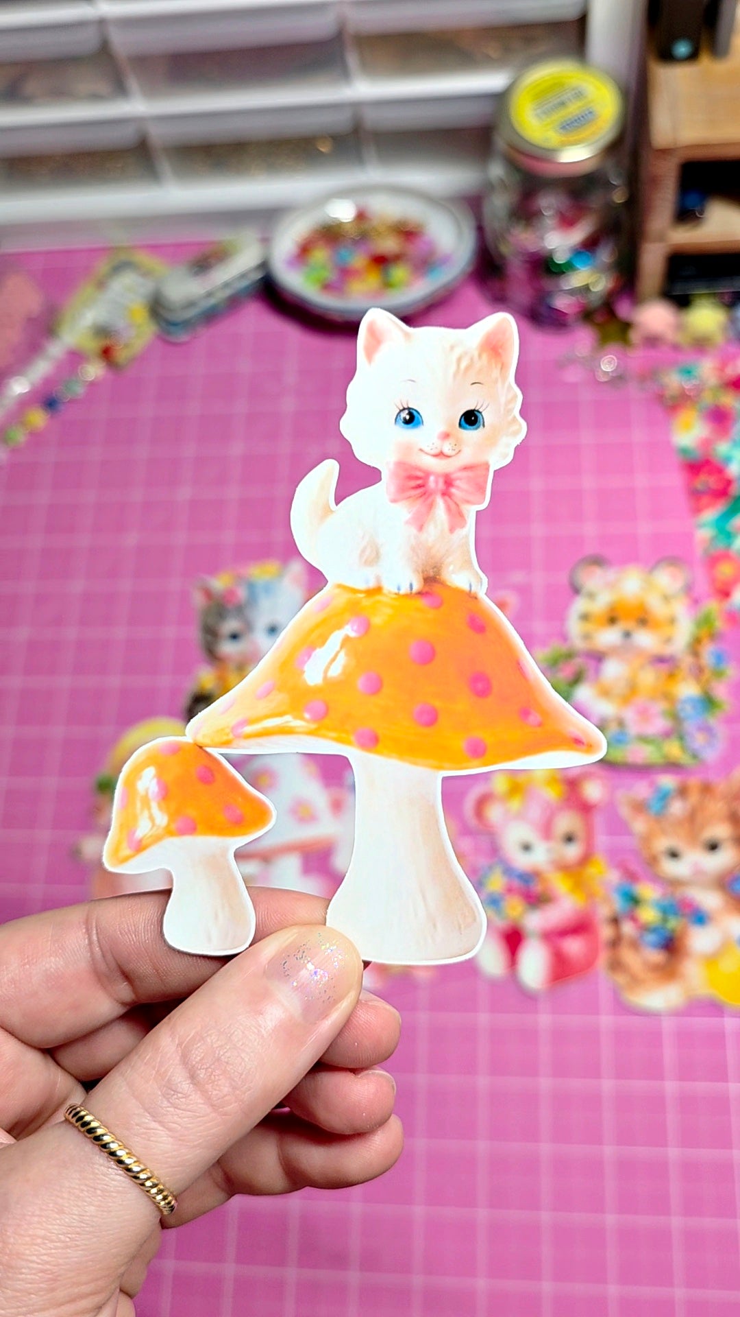 Cartoon cat on a mushroom with a pink bow held by a hand against a colorful background.