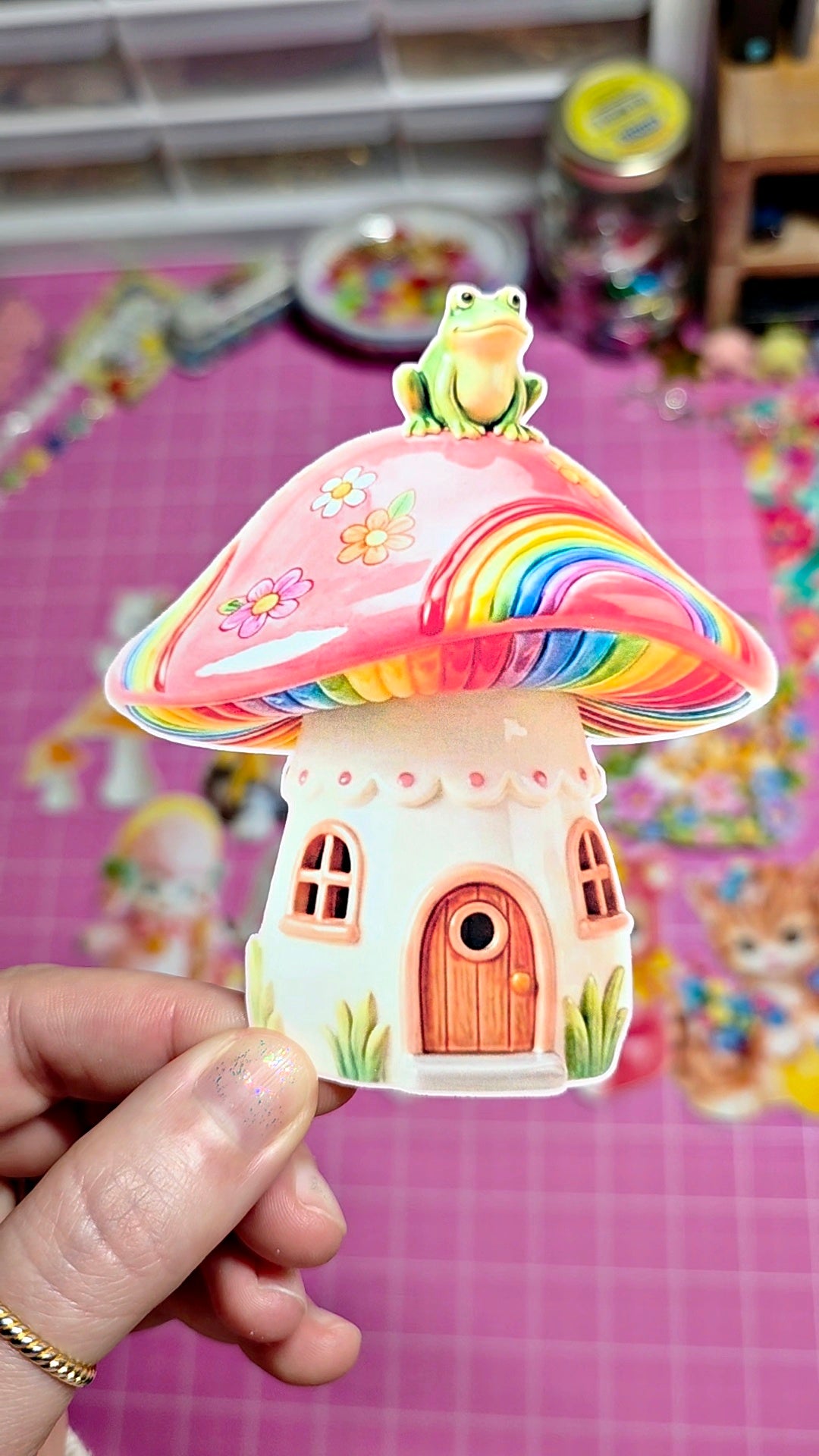 Colorful mushroom-shaped house with a frog on top, held by a hand against a colorful background.