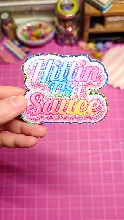 A hand holds a colorful, glittery die-cut sticker that reads 'Hittin' the Sauce' in pink, blue, and yellow lettering against a pink cutting mat background.