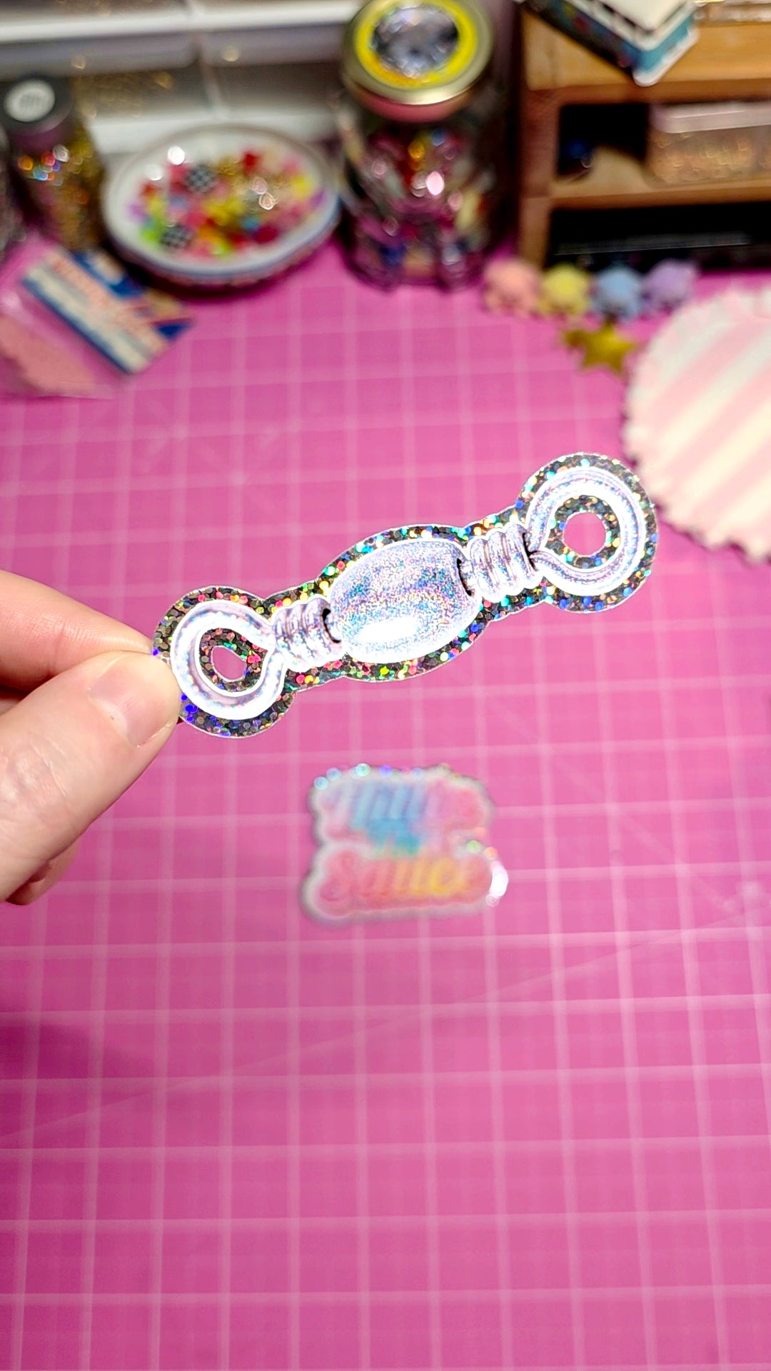A hand holds a small, iridescent, glittery key-shaped embellishment over a pink cutting mat, with a matching sticker or decal below it and craft supplies blurred in the background.