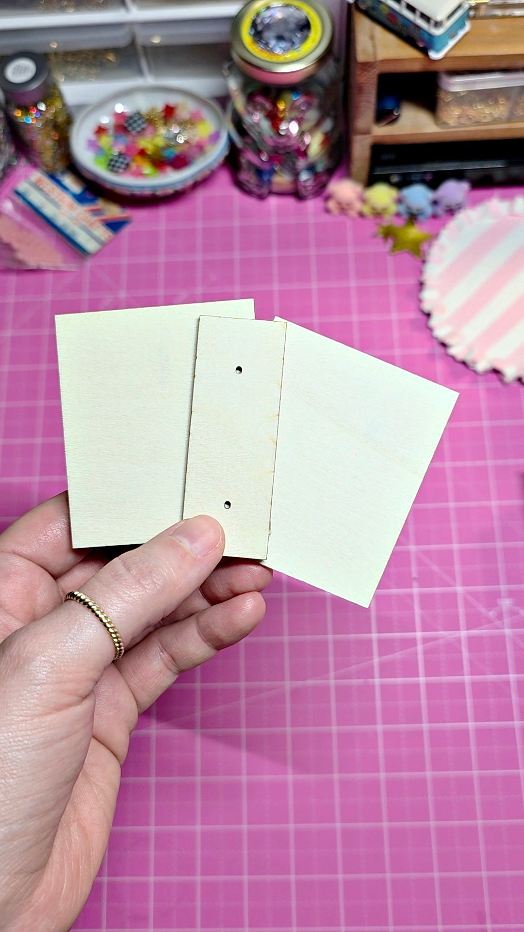 A hand holding three small cream-colored paper tabs or cards with pre-punched holes, set against a pink cutting mat with craft supplies in the background.