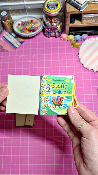A person's hands holding a small illustrated notebook or card set titled 'Scuffy' against a pink cutting mat background with craft supplies.