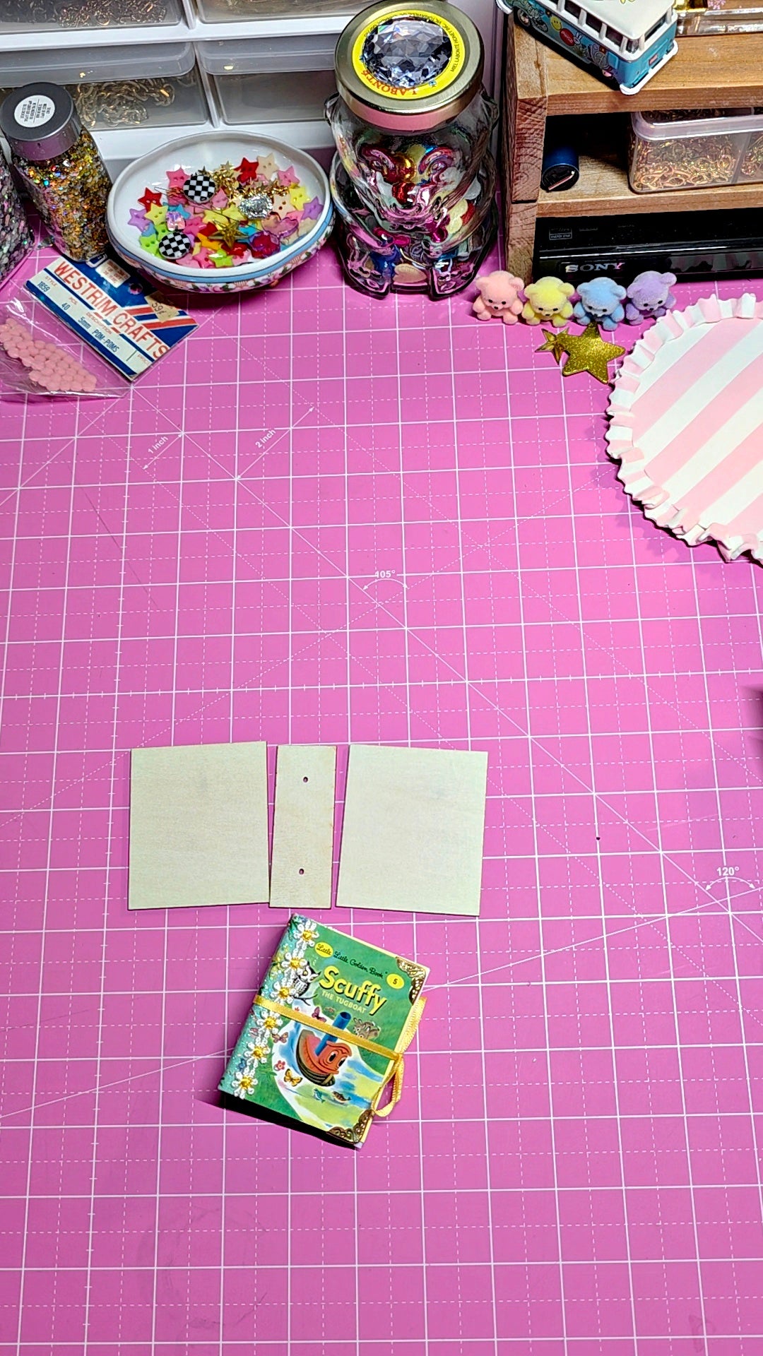 A pink cutting mat with a white grid pattern on a desk, featuring three blank beige cardstock sheets and a small illustrated notebook. The background is cluttered with craft supplies, including jars of glitter, a small wooden shelf, and a striped paper plate.