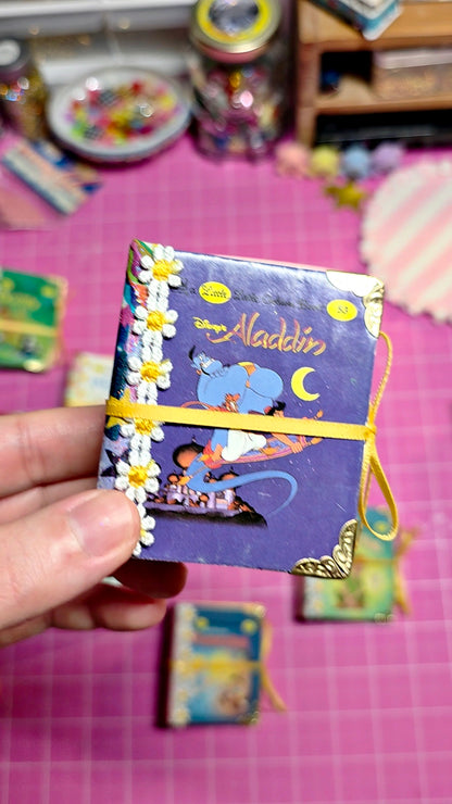 A hand holds a miniature purple Aladdin-themed notebook decorated with white daisy flowers and a yellow elastic band, set against a pink cutting mat background with other small items.