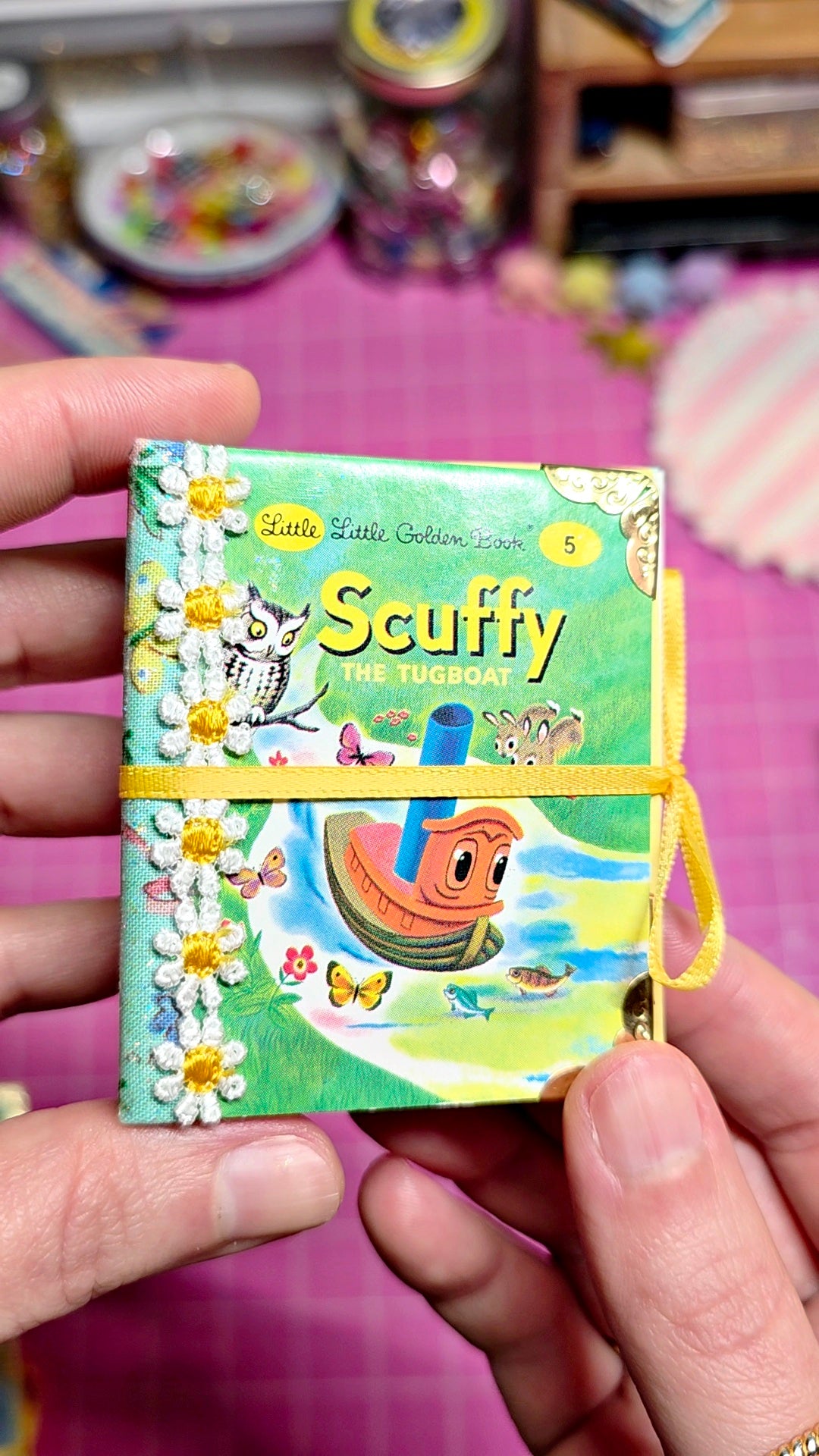 A person's hands holding a small illustrated book titled 'Scuffy the Pig' against a pink craft table background with other colorful items blurred in the distance.