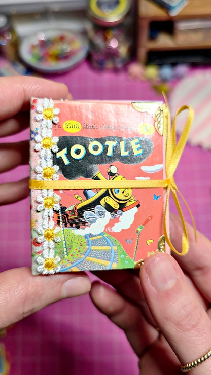 A person's hands holding a miniature Tootle toy book with a pink cover, yellow ribbon, and white daisy embellishments, set against a pink craft table background.