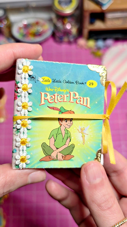 A hand holds a small decorative book titled "Peter Pan" with a yellow ribbon tied around it. The cover shows Peter Pan and Tinker Bell on a light blue-green background, with white daisy embellishments along the left edge.