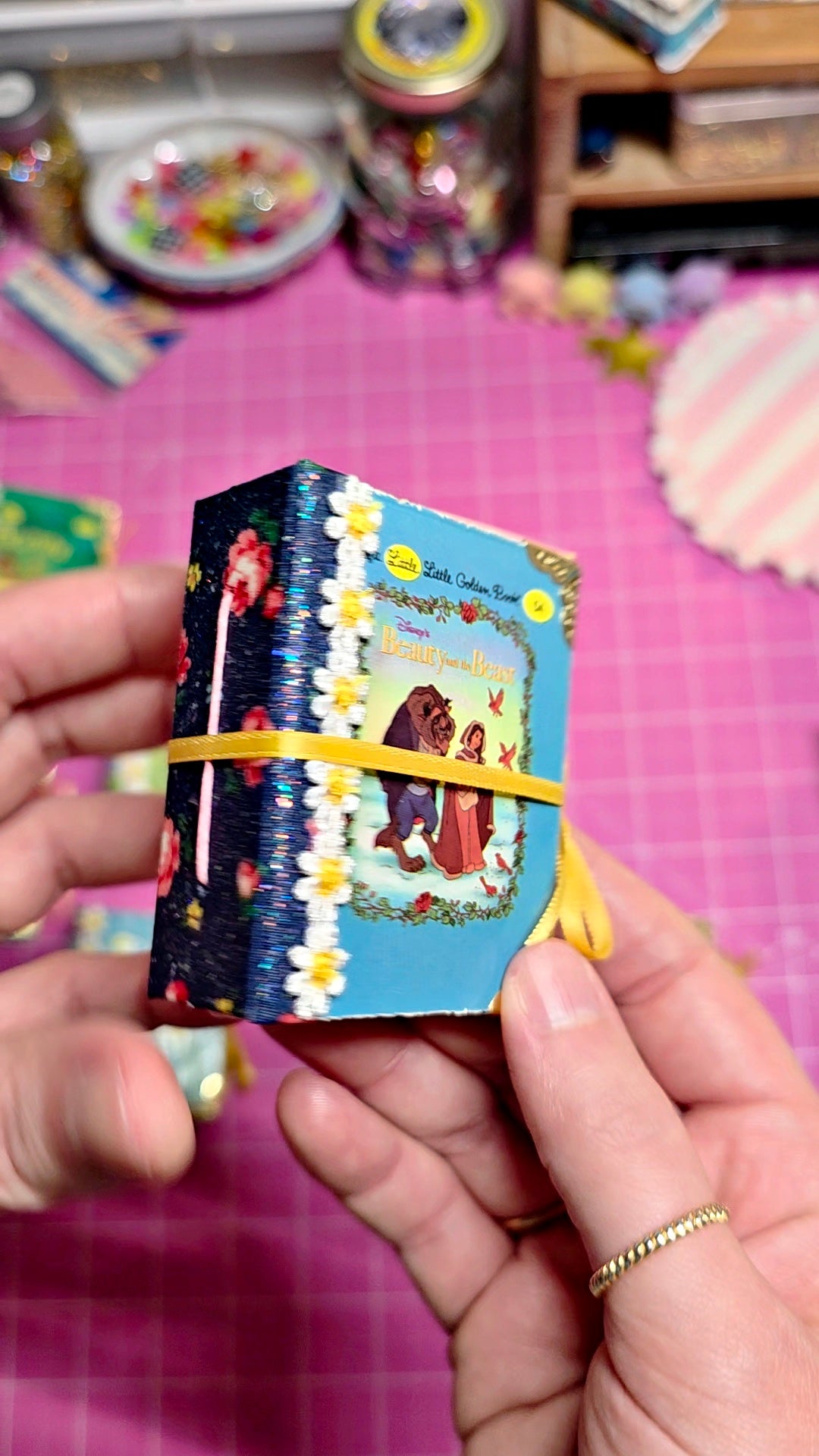 A miniature decorative book or journal wrapped with a yellow elastic band, held by two hands over a pink cutting mat background.