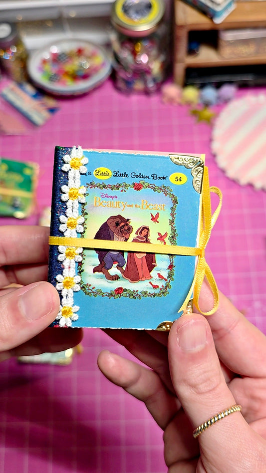 A miniature book journal featuring 'Little Little Golden Book' with a blue cover, white daisy decorations, and a yellow ribbon closure, held by hands over a pink cutting mat background.