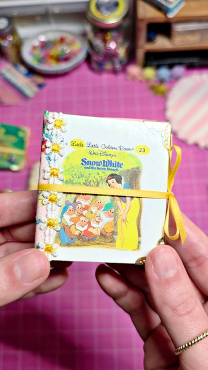 A hand holds a small illustrated notebook or card set featuring Snow White and the Seven Dwarfs, tied with a yellow ribbon, against a pink craft-table background.