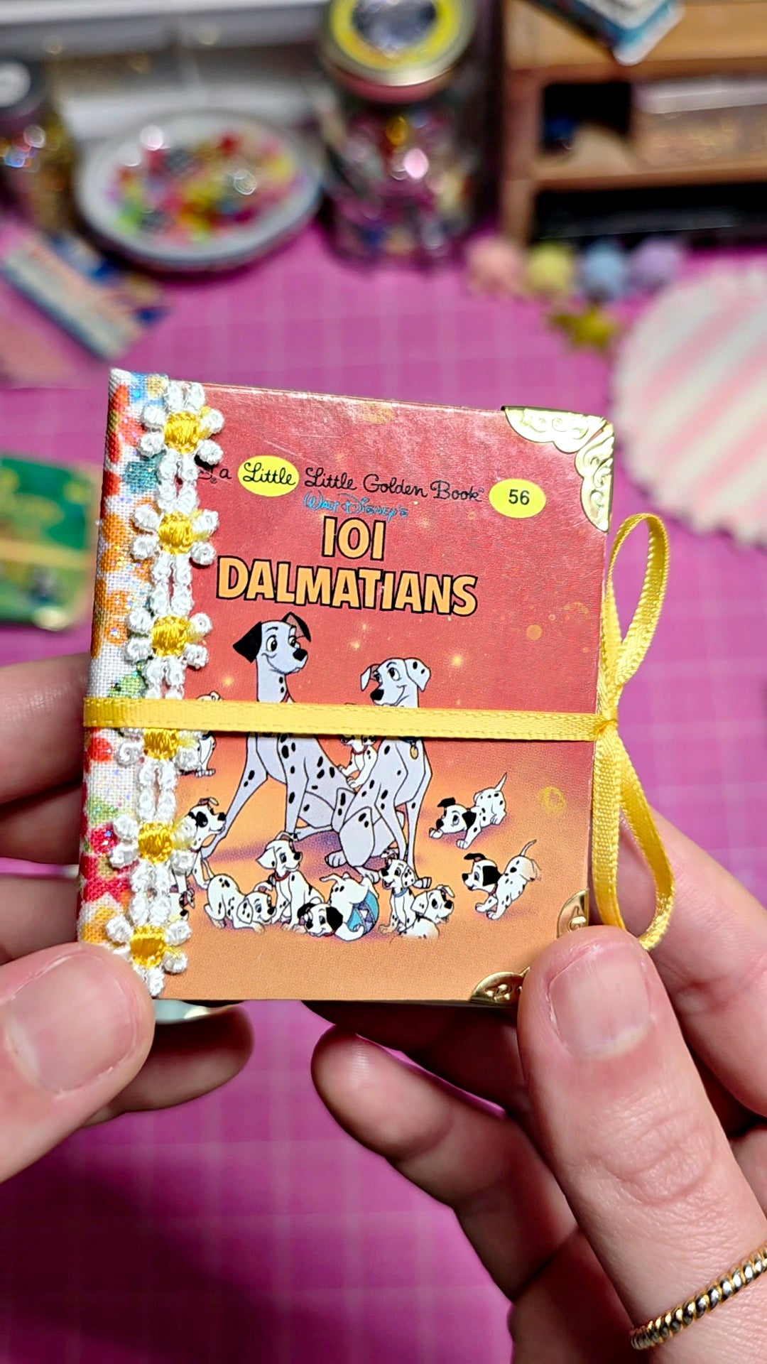 A hand holds a miniature hardcover book titled '101 Dalmatians' with a yellow ribbon loop, set against a pink cutting mat and craft supplies background.