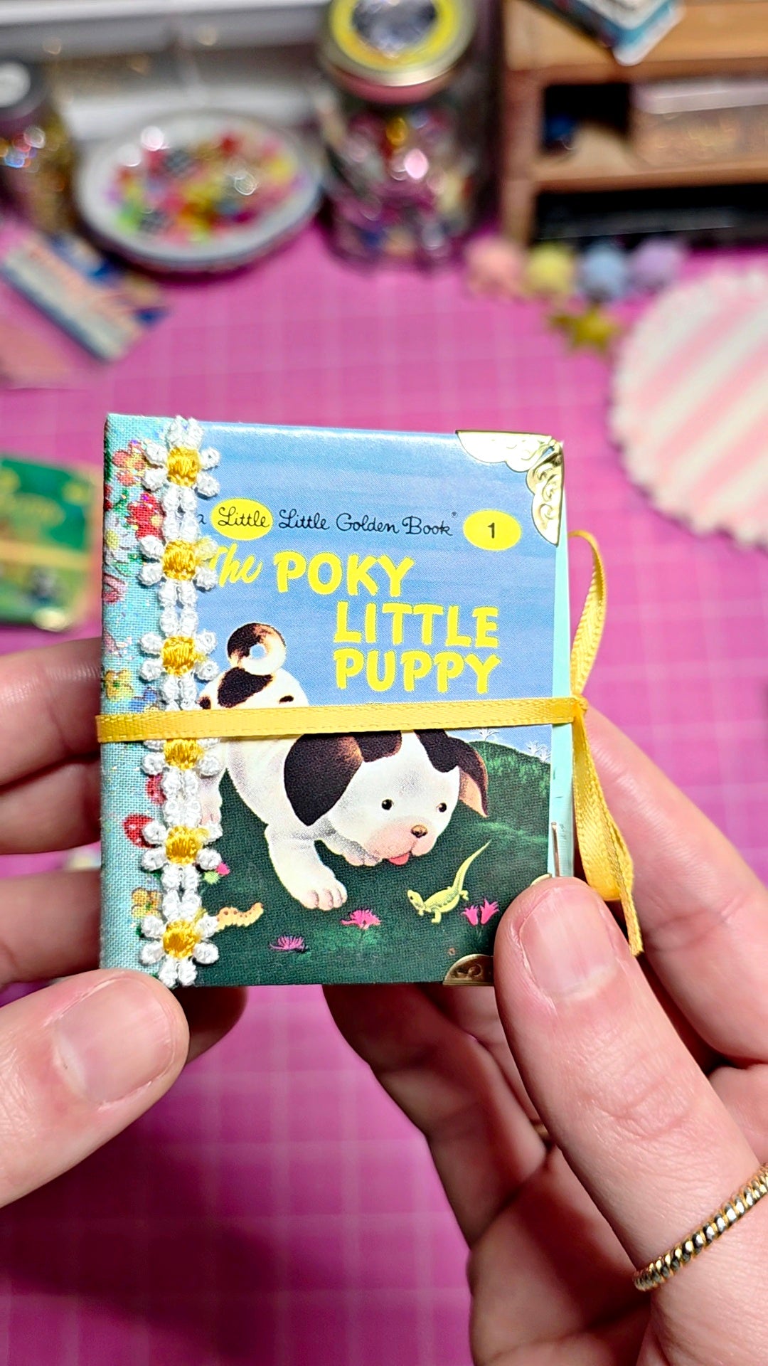 A miniature book titled "The Pokéy Little Puppy" held in two hands. The book is decorated with a yellow ribbon, white daisy embellishments, and a printed illustration of a black-and-white puppy on a blue and green background.