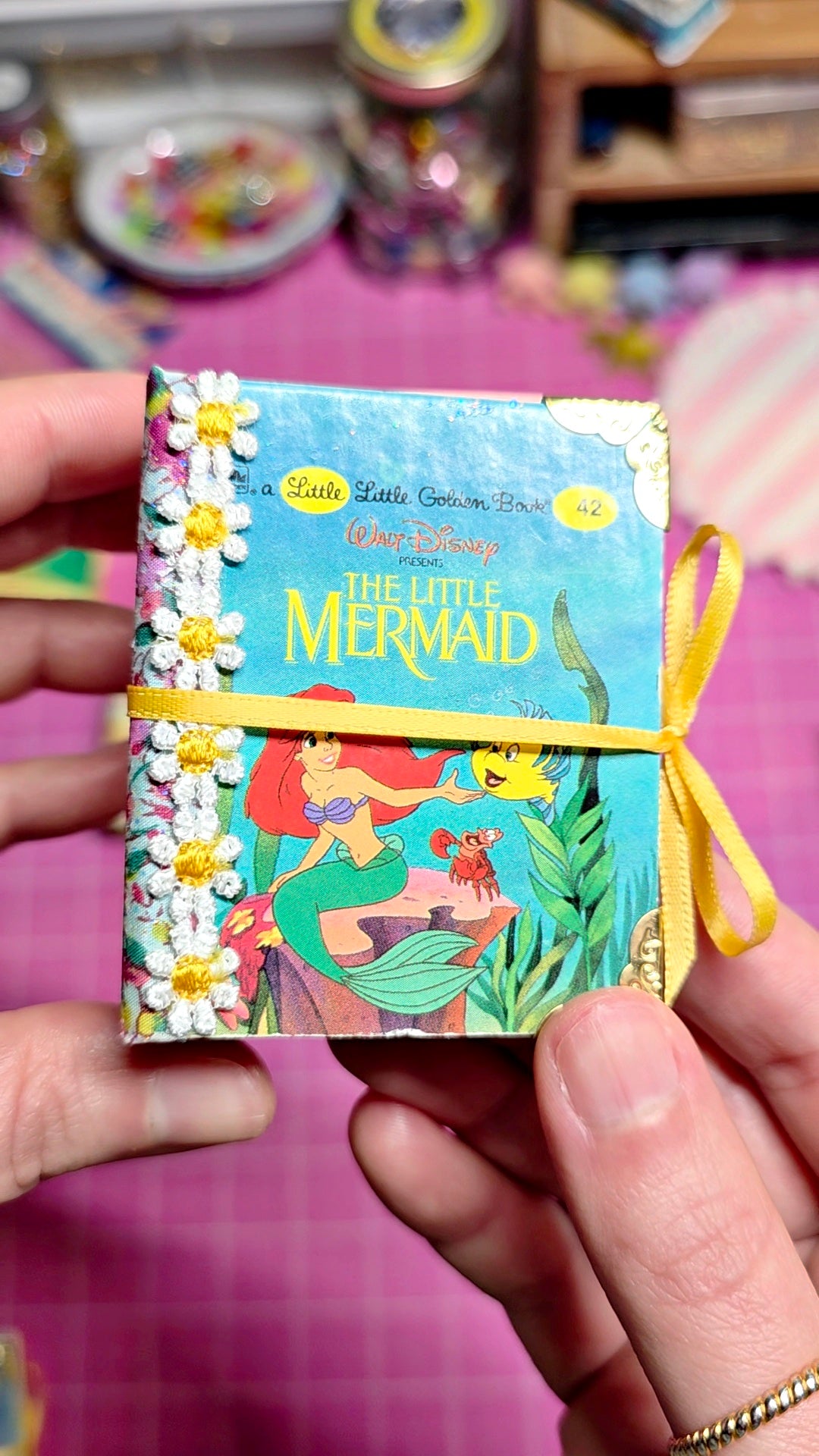 A person holds a small Disney 'The Little Mermaid' book or notebook with a colorful mermaid illustration, white daisy flowers along the spine, and a yellow ribbon tied around it, against a pink background with blurred shelves.