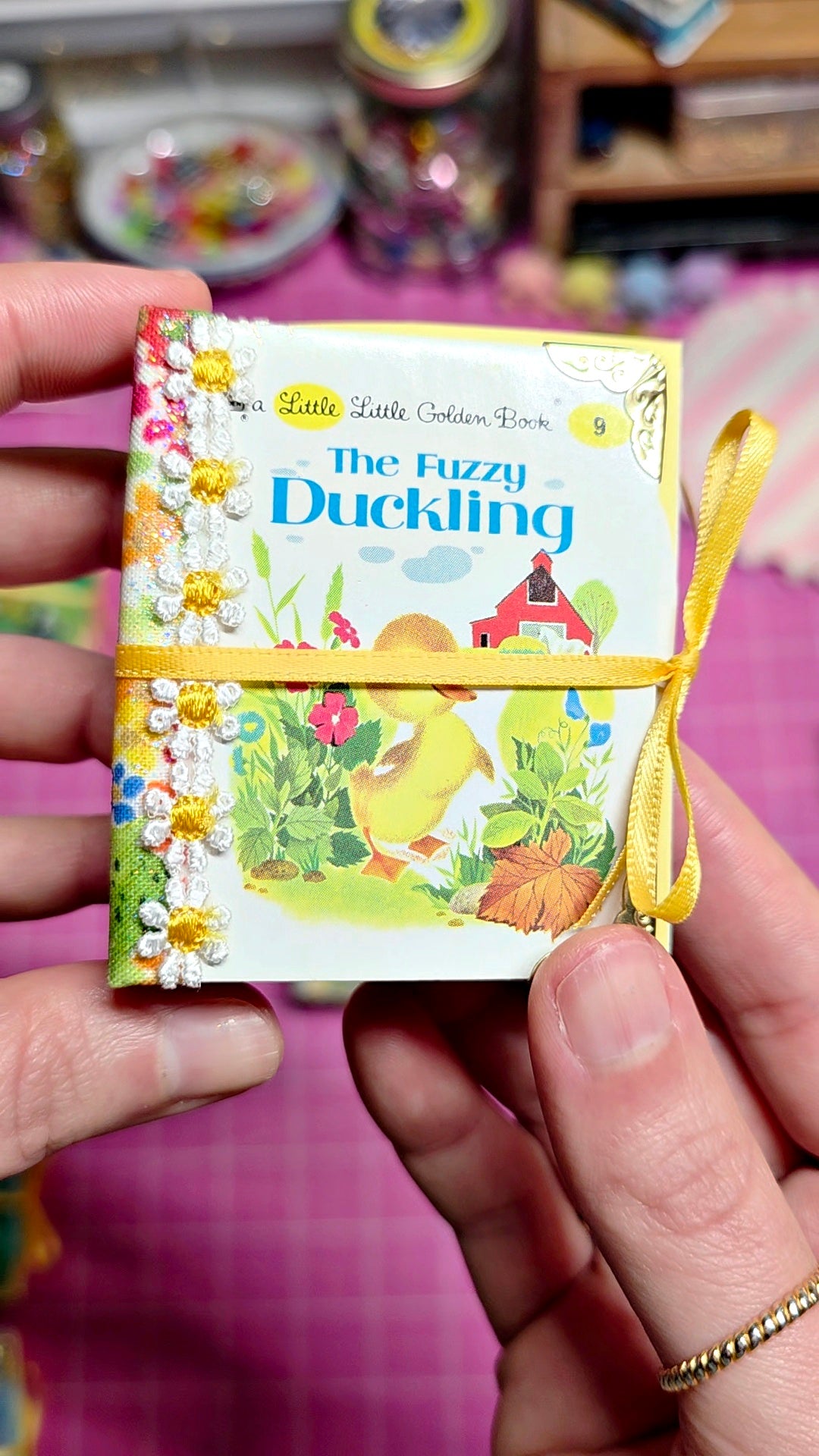 A hand holds a small illustrated book titled "The Fuzzy Duckling" with a yellow ribbon tied around it. The cover shows colorful duckling and floral artwork, and the background is a pink table with blurred craft supplies.